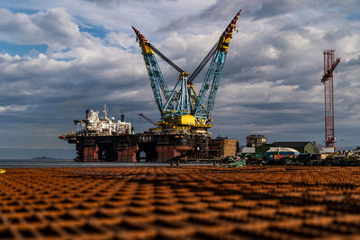 Cranes at oil rig in sea against cloudy sky
