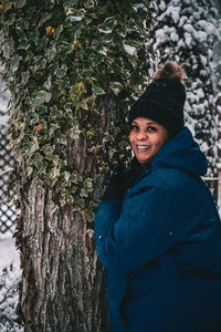 Portrait of smiling young woman standing by tree trunk during winter