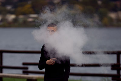 Teenage boy smoking while holding electronic cigarette against lake