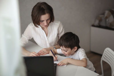 Beautiful mother teaches her son at home at the table, real interior