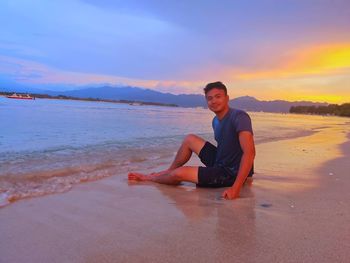 Portrait of man sitting on shore at beach against sky during sunset