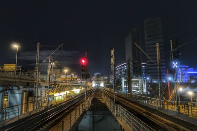 Illuminated railroad tracks amidst buildings against sky at night