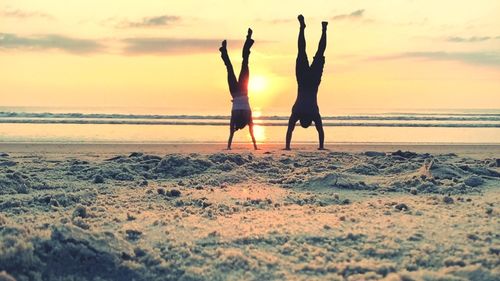 Full length of friends doing handstand at sandy beach against orange sky