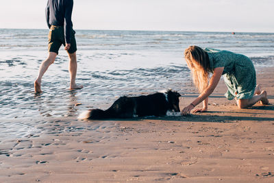 Full length of young woman feeding dog with unknown person on beach