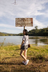 Side view of young woman standing by signboard on field against lake in forest