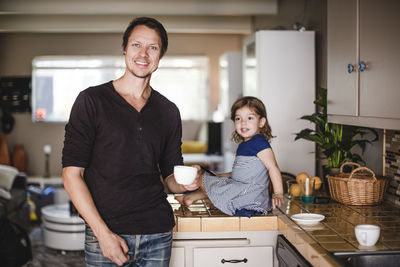Portrait of smiling mature man with cup while daughter sitting on kitchen counter