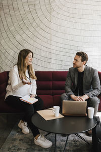 Pregnant businesswoman gesturing while discussing with male professional sitting on sofa at convention center