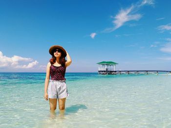 Full length portrait of young woman standing at beach against sky