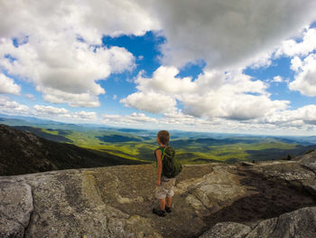 Rear view of boy standing on rocky mountain peak while looking at landscape against sky