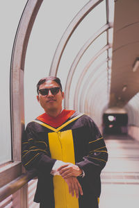 Portrait of young man wearing sunglasses and graduation gown while standing in corridor