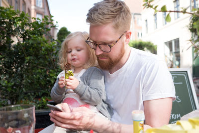 Father using smart phone while sitting with daughter holding chocolate at sidewalk cafe