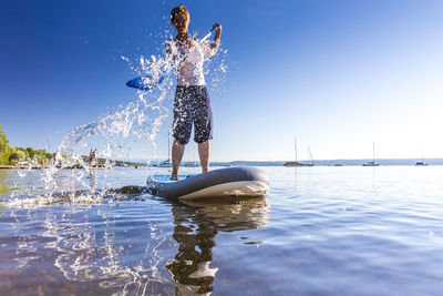 Full length of young woman paddleboarding in lake against clear blue sky