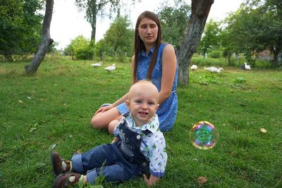 Portrait of boy with mother sitting by bubble on grassy field at park