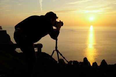 Silhouette man photographing sea against sky during sunset