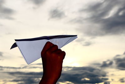 Cropped hand of girl flying paper airplane against sky
