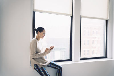 Woman using mobile phone while sitting on window sill at home