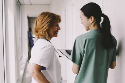 Happy female healthcare workers looking at each other while standing in hospital corridor