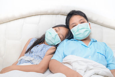 Brother and sister wearing medical face mask sitting on bed at home, 