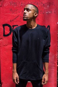 Young man looking away while standing against red wall