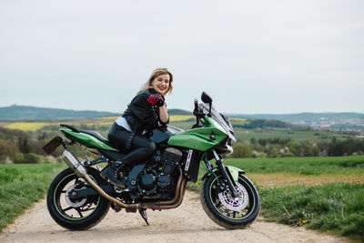 A woman in a leather jacket poses on a green motorcycle parked on a dirt road. the scene conveys a