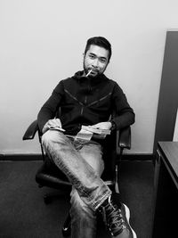 Portrait of man with cigarette in mouth holding currency bundle while sitting on chair against wall