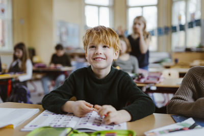 Smiling male pupil with blond hair sitting at desk in classroom