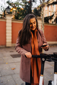 Hispanic long haired lady in warm coat standing on city street and doing contactless payment for electric scooter with smartphone using application