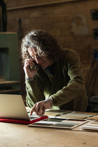 Concentrated male carpenter having phone call on mobile phone while typing order on netbook during work in joinery