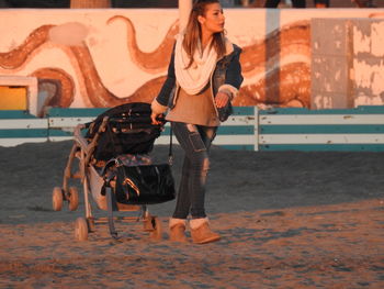 Young woman pulling baby stroller while walking on sand against graffiti wall