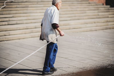 Full length of senior man cleaning street using garden hose in city