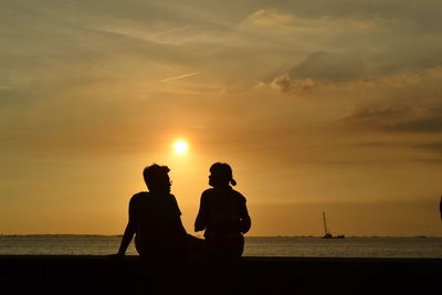 Silhouette men sitting on shore against sunset sky