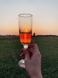 Midsection of person holding drink on field against sky during sunset