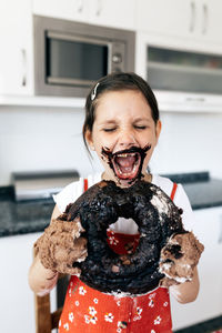 Child with dirty face and hands standing with tasty ring cake and open mouth at table in house kitchen