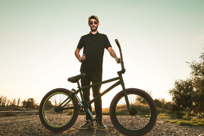 Young man with bicycle standing on field against clear sky during sunset