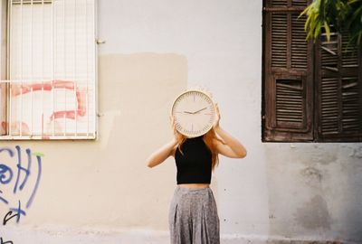Woman holding clock while standing against wall