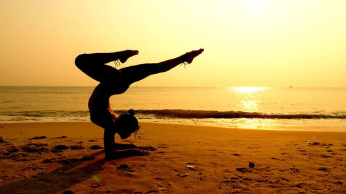 Full length of woman practicing yoga on shore at beach against sky during sunset