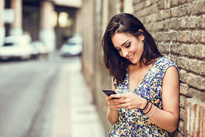 Smiling young woman using mobile phone while leaning on brick wall