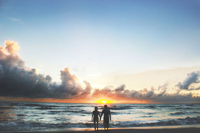 Rear view of friends standing on beach against sky during sunset