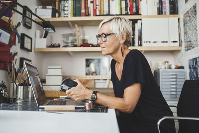 Female industrial designer using laptop while holding product at home office