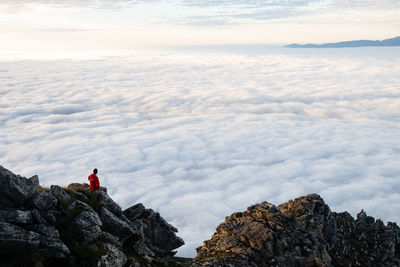 Man in red jacket sitting on cliff rock looking at the sea of cloud below 