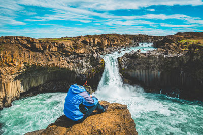 Rear view of man sitting on rock looking at waterfall