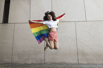 Cheerful woman jumping while holding rainbow flag in front of wall