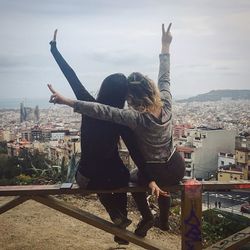 Rear view of female friends with arms raised sitting on railing overlooking cityscape against sky