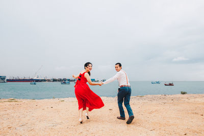 Rear view of couple holding hands while standing on beach against sky