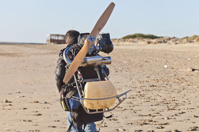 Rear view of boy with equipment at beach against clear sky