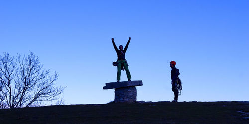 Male hiker by friend with arms raised standing on rock at mountain against clear blue sky