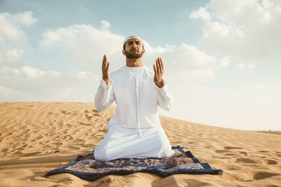 Man praying on sand at desert against sky
