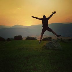 Full length of man jumping on land against mountains and sky during sunset