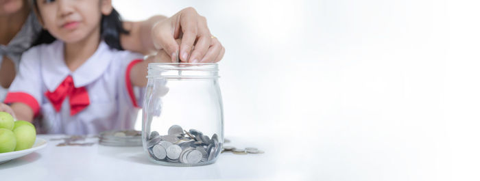 Cropped hand of mother with daughter putting coin in jar against white background