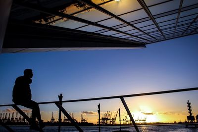 Silhouette man sitting by river on railing during sunset